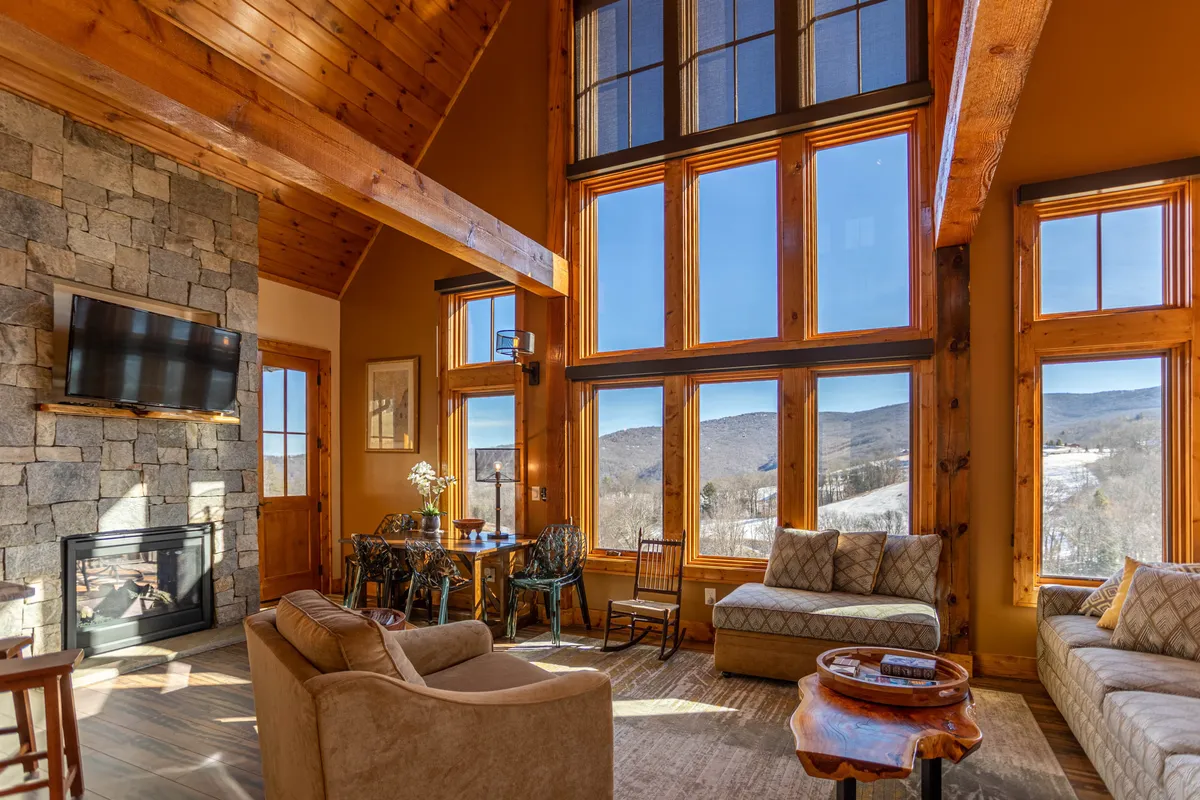 A welcoming living area with large windows showcasing a mountain view, a striking stone fireplace, wooden beams, and comfortable seating arranged for relaxation and conversation. The ambiance is inviting and serene, perfect for family gatherings or quiet evenings.