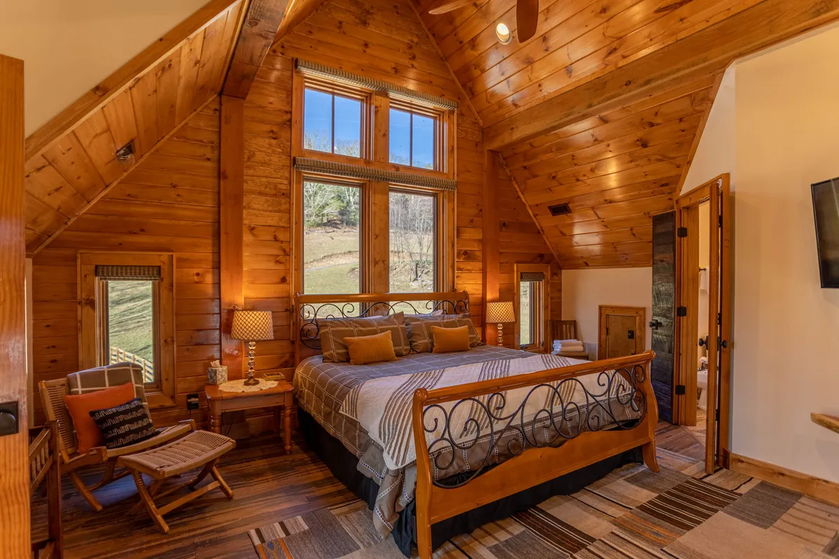 A spacious bedroom with a plush king bed, large windows offering a beautiful view, a wooden ceiling, and decorative lamps. The space feels tranquil and warm, perfect for relaxing or reading by the window.