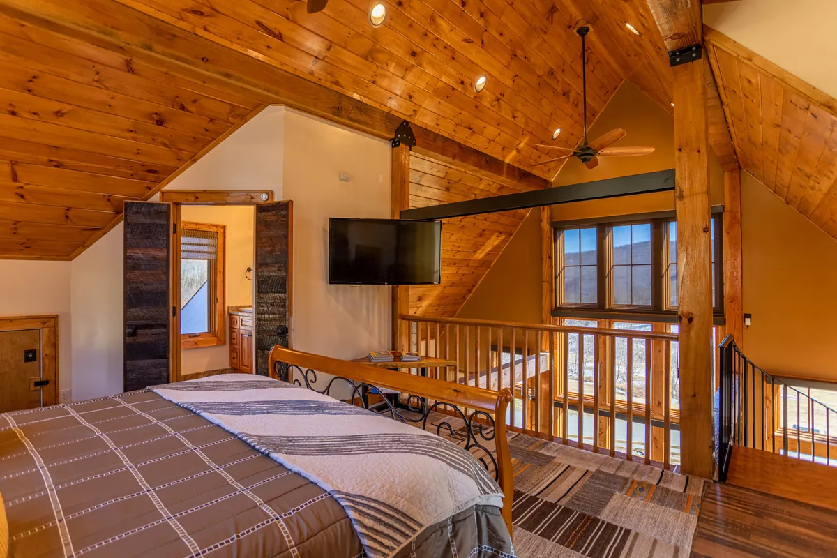 A cozy bedroom with a bed featuring patterned bedding, large windows letting in natural light, a TV mounted on the wall, and a wooden railing. The space is warm and welcoming, ideal for relaxing or enjoying TV in bed.