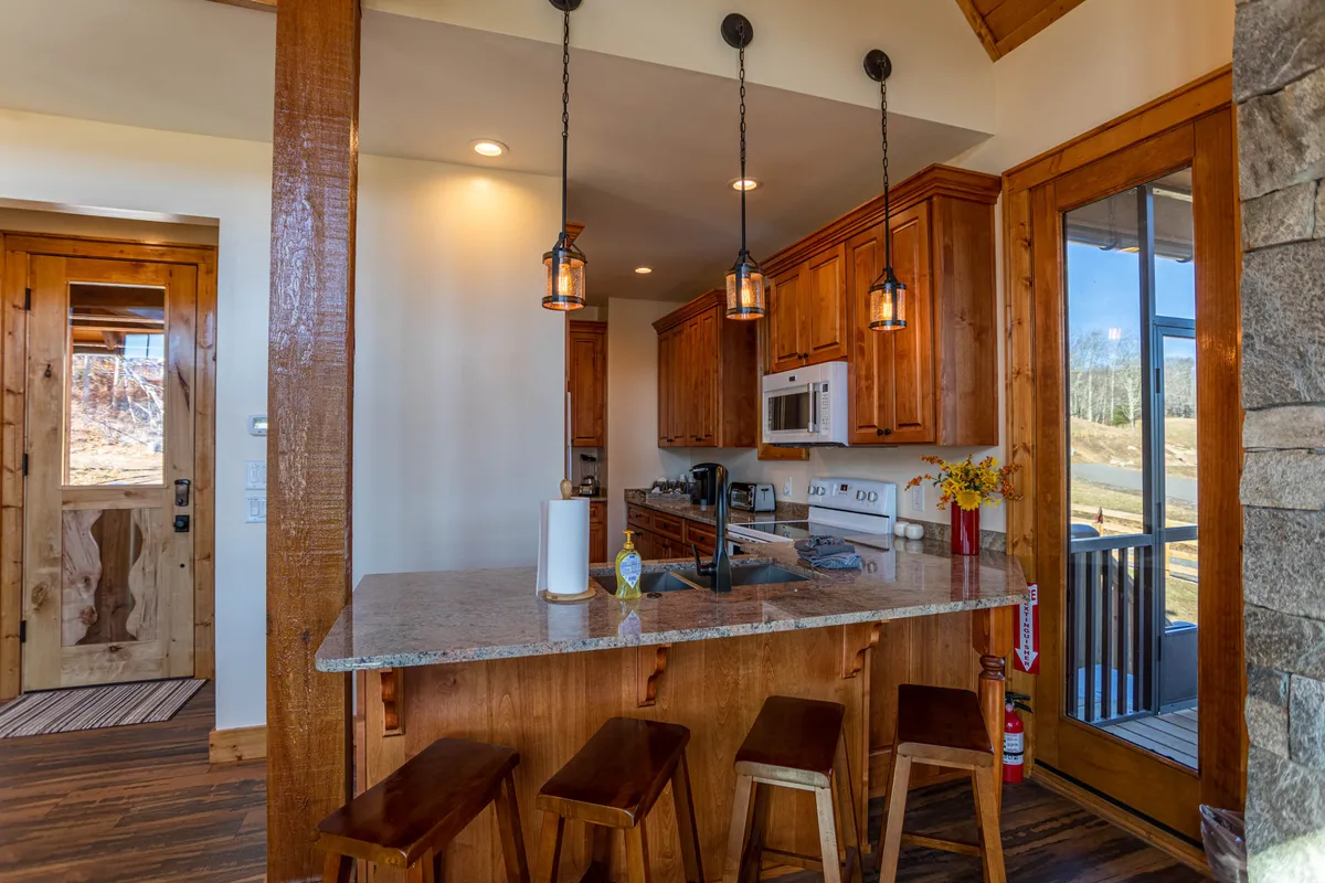 A kitchen scene with a granite countertop, warm wooden cabinetry, modern pendant lighting, and a view of the outdoors. Designed to encourage cooking and socializing, the kitchen is both functional and visually appealing.