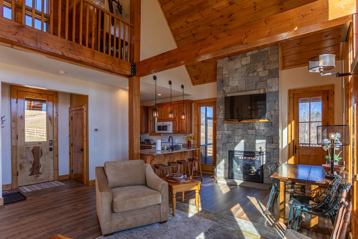 This image displays a cozy, open-concept living space featuring a fireplace, large windows that fill the room with natural light, exposed wooden beams, a modern kitchen area, and a dining table. The scene exemplifies the integration of living, dining, and kitchen spaces in a welcoming, communal atmosphere.