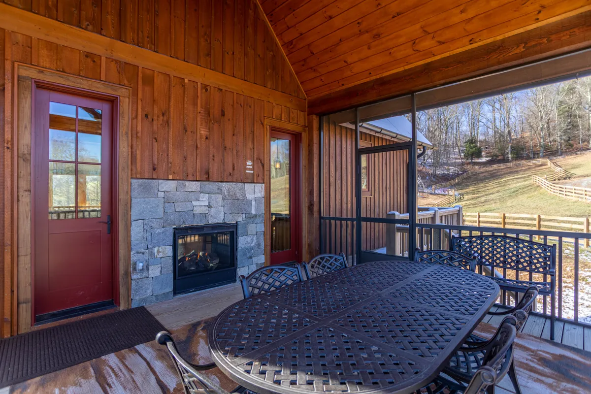This image presents an inviting outdoor dining area with a sturdy wooden table, backed by a cozy outdoor fireplace set into the wooden exterior walls. Large windows offer glimpses of the scenic outdoors, while the table is ideally positioned for gatherings and meals. The space exudes a warm, welcoming mood, perfect for morning coffee or evening feasts.