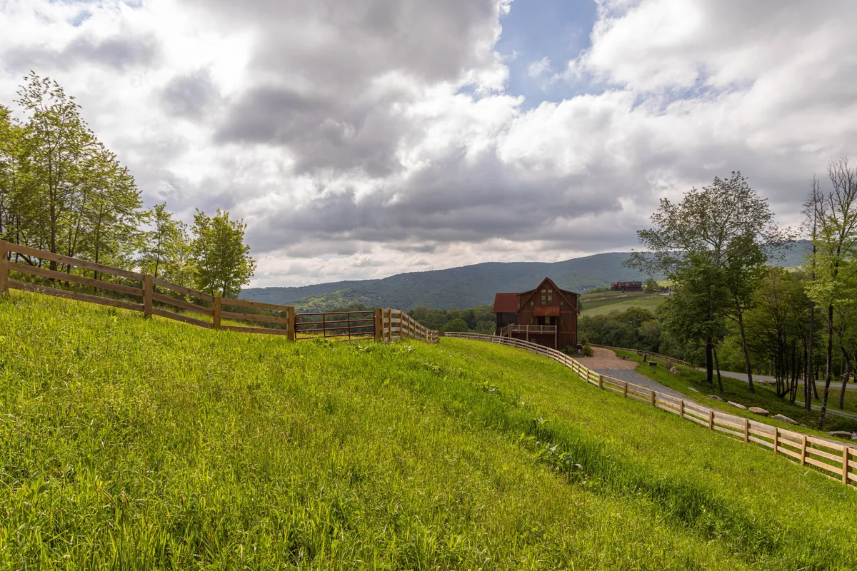 An exterior shot with an expansive green lawn, wooden fence, and mountain backdrop. While not showing horses directly, the fenced landscape and rustic feel evoke the property's equestrian spirit and hint at the presence of pastures and horse-friendly infrastructure.