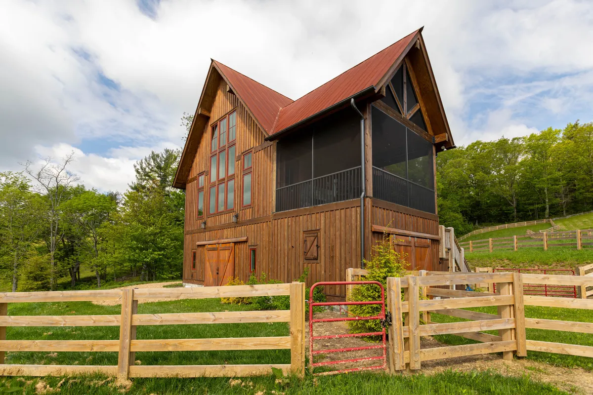A wide view of the barn-style house with striking architecture, large windows, and a prominent red gate. The surrounding landscape is rustic and open, suggesting ample space for horses and direct access to outdoor activities and trails.