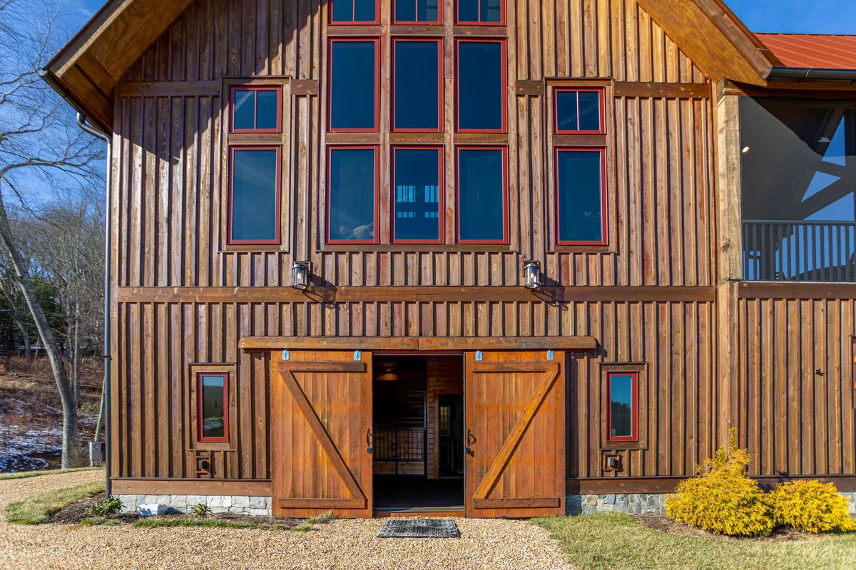 The image features the rustic facade of the home with large wooden doors, tall windows, and red accents, set against a natural backdrop. The inviting entrance and outdoor space hint at shared gatherings, wine evenings on the porch, and an overall sense of welcoming luxury.
