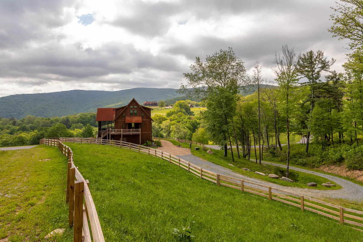 This image showcases the rustic wooden house nestled among lush greenery, with a winding driveway leading up to the home and mountains visible in the distant background. The natural landscape invokes a sense of serenity and connection to the outdoors, perfectly fitting the narrative of a luxurious mountain escape and scenic relaxation.