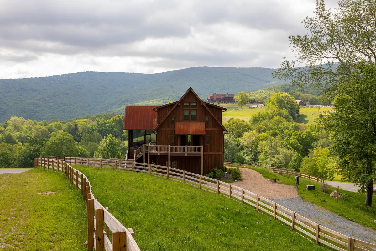 A rustic wooden house nestled beside a fenced pathway, with the surrounding mountains visible in the background. The area is surrounded by greenery, inviting families to explore, gather outdoors, and enjoy peaceful natural vistas both at sunrise and sunset.