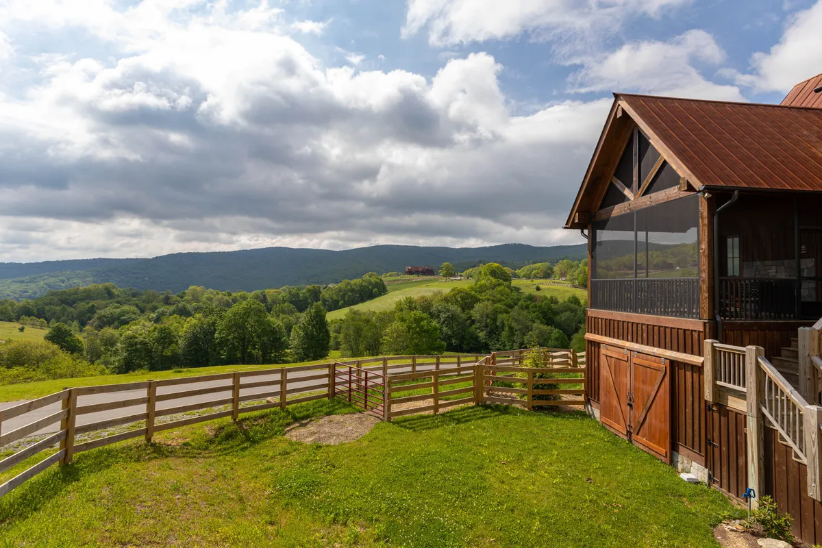 The image features a stunning wooden cabin perched amid rolling green hills, framed by a rustic fence and set beneath a peaceful, cloudy North Carolina sky. The surrounding landscape is open and inviting, ideal for outdoor activities like hiking or simply soaking in panoramic mountain views. The serene setting promises seclusion and a close connection to nature, with the cabin's exterior suggesting warmth and modern comfort.