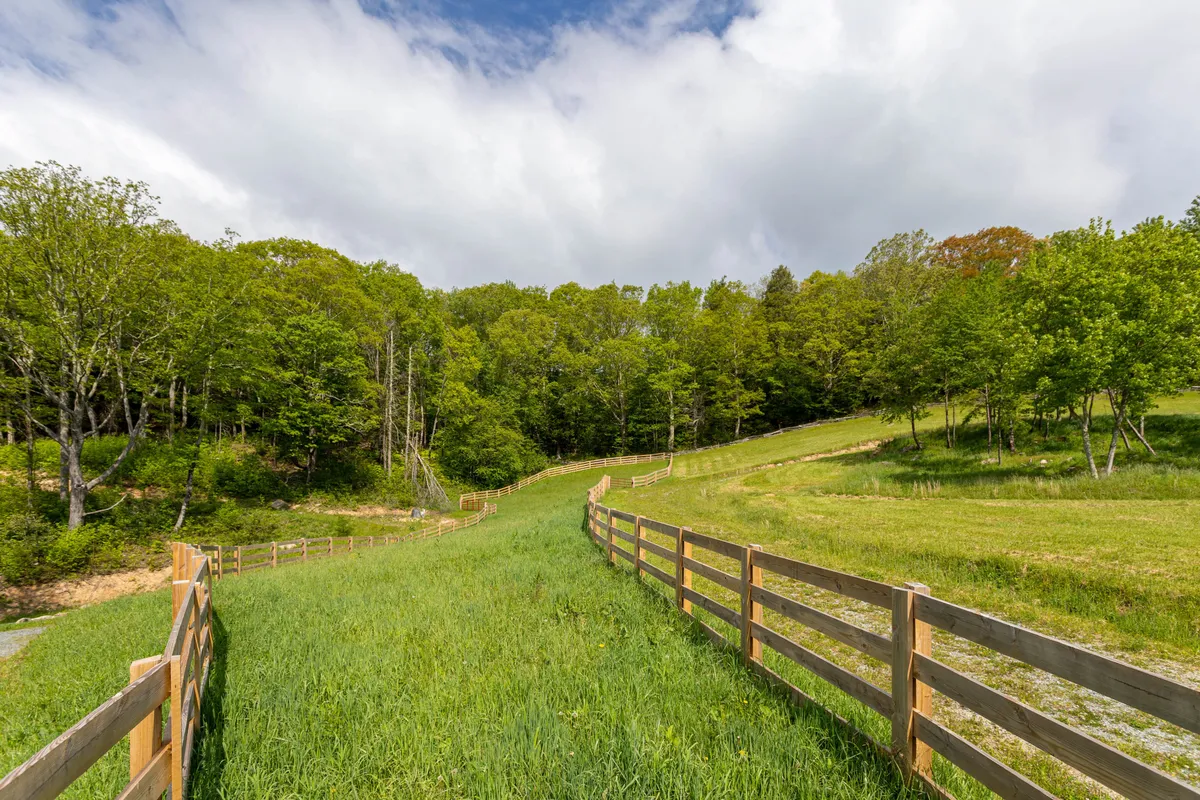 This exterior image showcases a wooden fence enclosing a lush green pasture with a backdrop of tall trees. The open, grassy area evokes a peaceful, rural setting ideal for horse-related activities and pasture rotation. The fence is a prominent feature, underscoring the property's equestrian-friendly infrastructure.