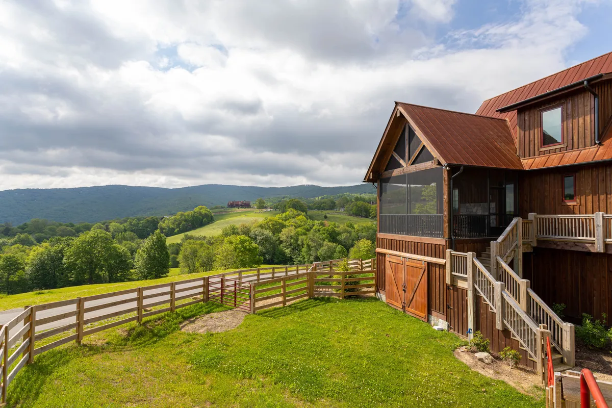 This image captures the house facade bordered by a sturdy fence, with rolling landscape and a visible stretch of road. The inviting porch and expansive yard communicate tranquility and a seamless indoor-outdoor connection, perfect after a day on the trails.