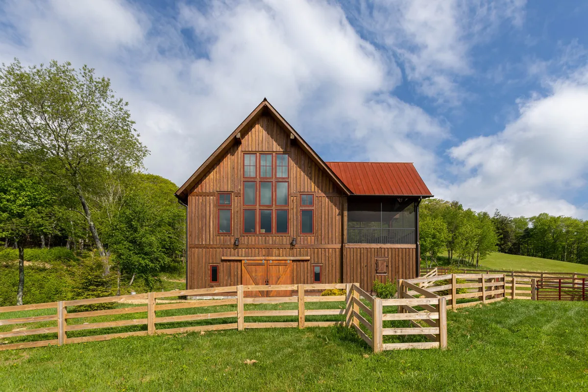 This image presents the exterior of the property with large windows, a red roof, and a fenced yard framed by surrounding greenery. The setting is serene and inviting, suggesting ample opportunities for outdoor gatherings, peaceful reflection, or enjoying a sunset after a day spent at the vineyard.